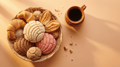 Assorted Mexican pan dulce sweet breads including conchas, cuernos, and orejas in a woven basket with a clay mug of coffee