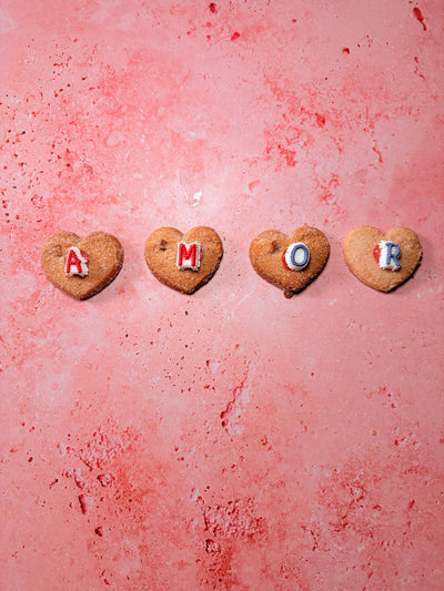 4 heart-shaped cookies in a row with a letter in each spelling out "AMOR" and pink background