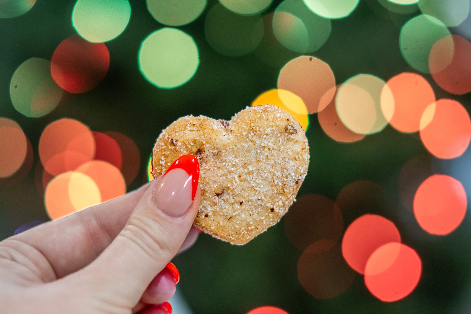 Heart-shaped cookie held up by two fingers with lights in background