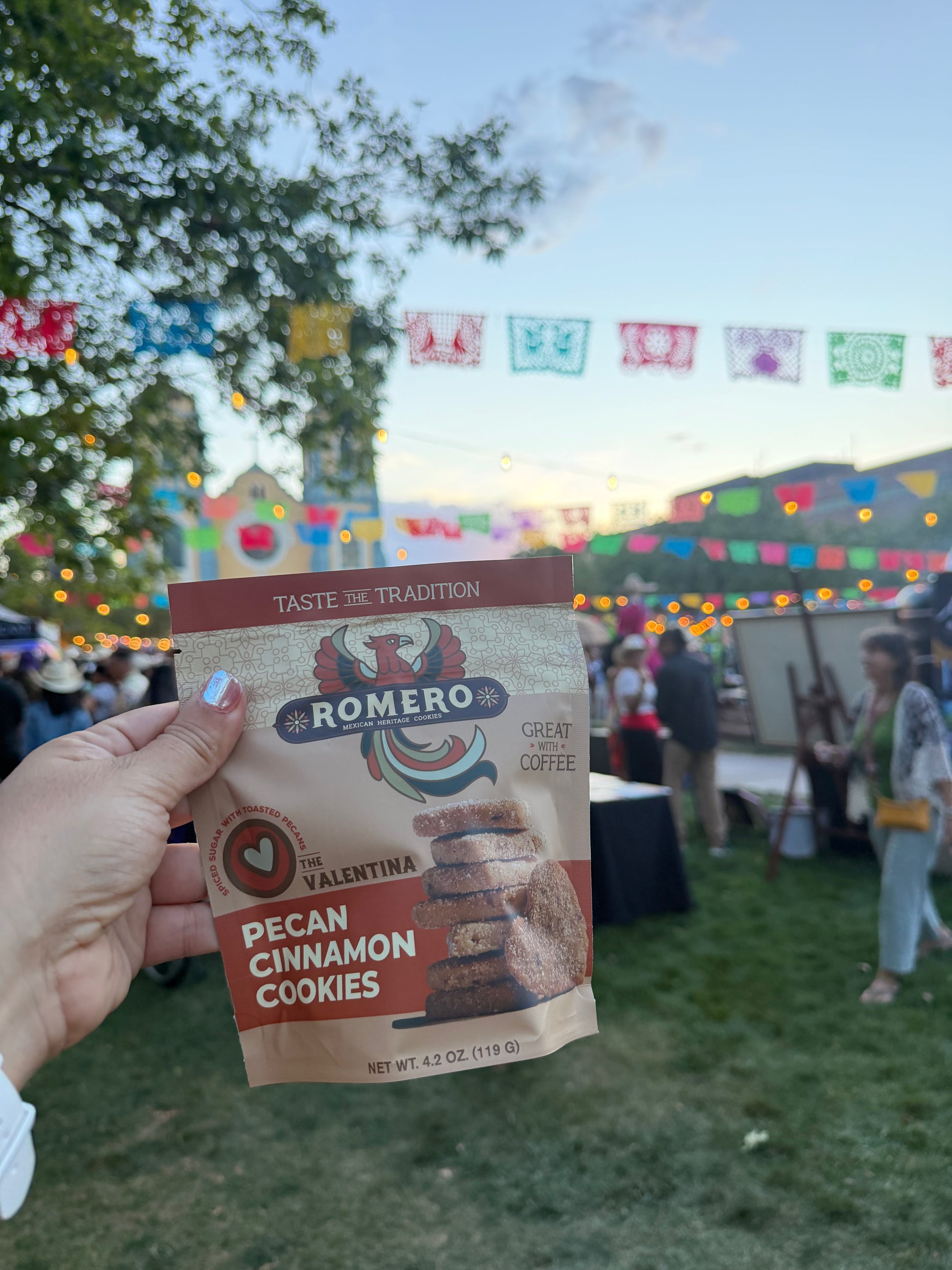 A hand holds a bag of Romero de la Valentina Pecan Cinnamon Cookies in front of an outdoor festival scene at dusk with colorful flags and string lights.