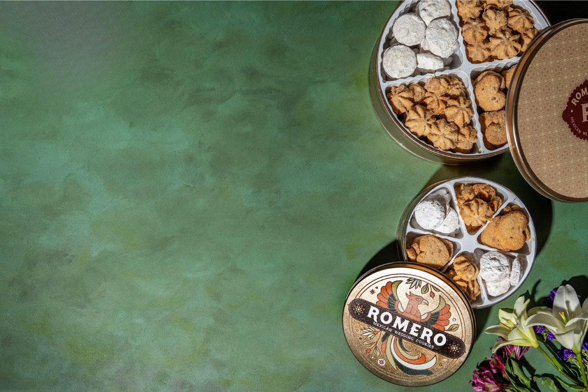 Two round metal gift tins filled with assorted cookies and chocolates, arranged on a textured green background with decorative flowers in the lower right corner. One tin lid is clearly visible and shows the brand name 'ROMERO'.