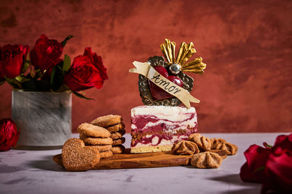 Decorative cake slice with 'Amor' sign, cookies, and red roses on a wooden board against a red background.