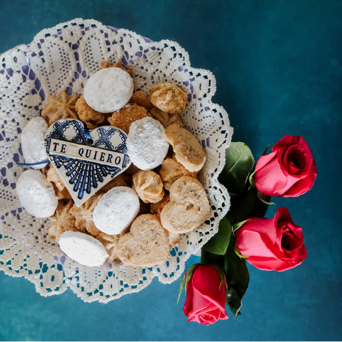 A heart-shaped cookie decorated with blue lace-like pattern and the words 'TE QUIERO' sits on a white lace doily with assorted cookies and powdered sugar cookies, next to three red roses on a dark teal background.