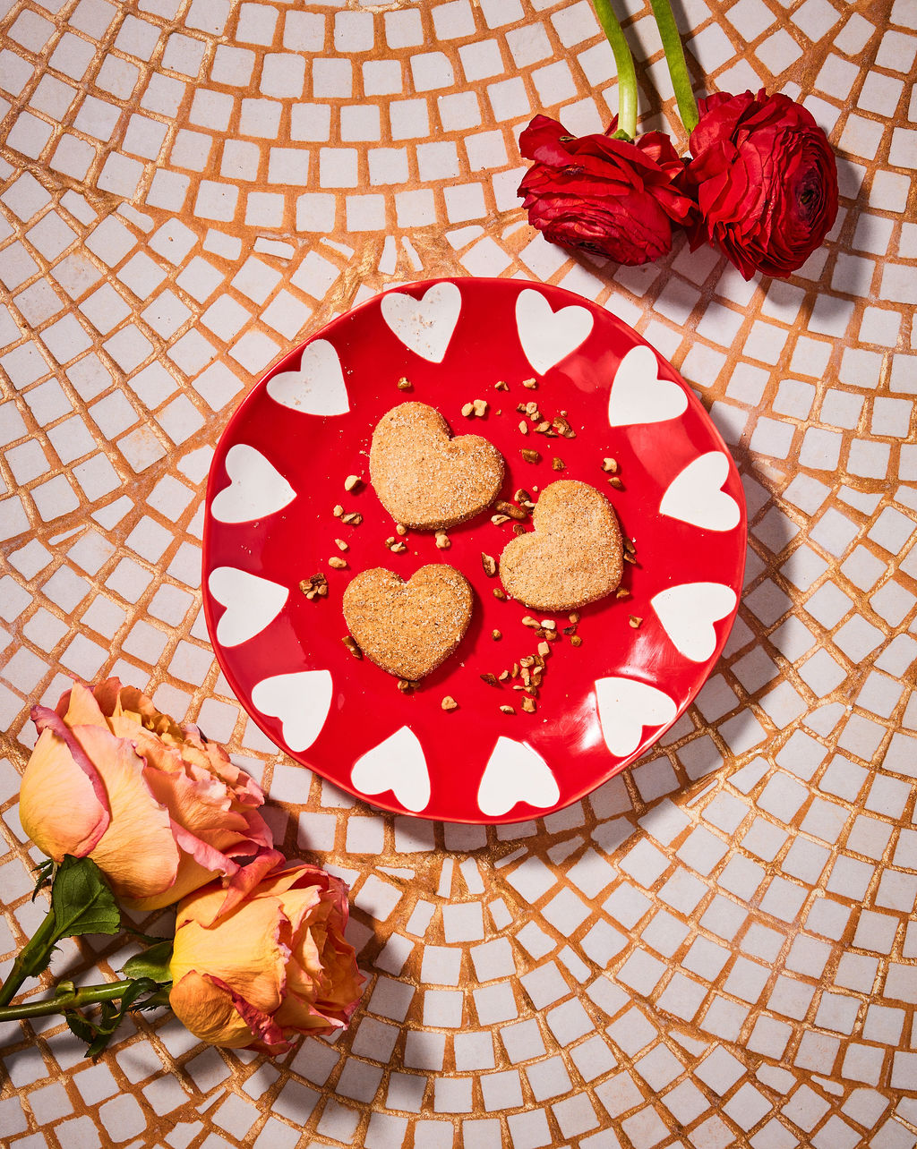 Heart-shaped cookies on a red plate with white hearts, surrounded by flowers on a mosaic tile background.